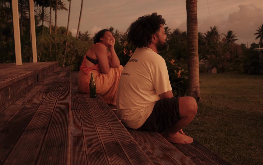 A man and a woman sit on the edge of a deck with beers in the last light of the day, the setting is in Samoa