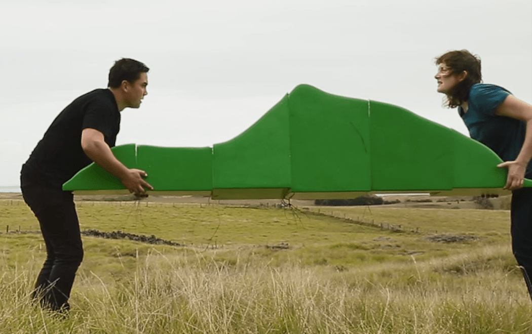 Rebecca Hobbs and Martin Awa Clarke Langdon hold a mountain like green sculpture between them whilst standing in a grassy field