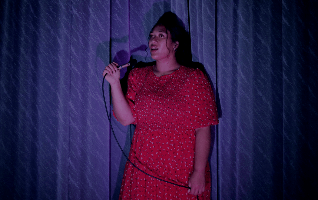 Ash Ulutupu wears a red dress sings alone into a microphone  in front of a blue stage curtain