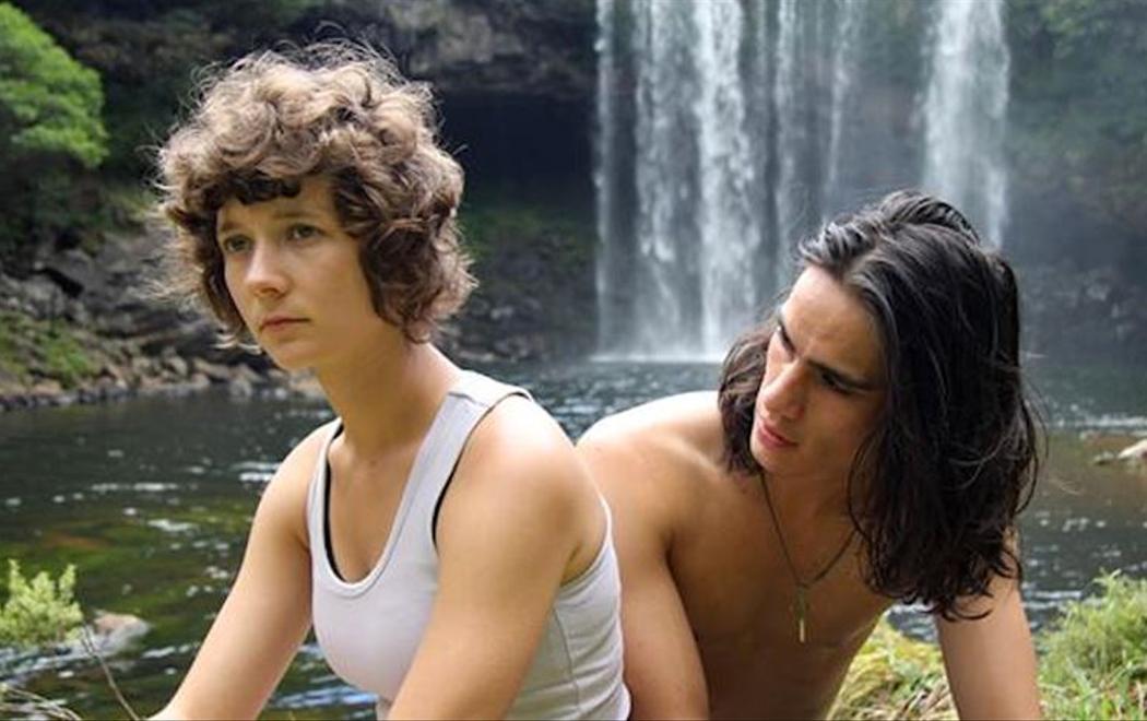 A woman stares off to the distance whilst a man next to her looks at her face, they sit by a pond with a waterfall in the background.