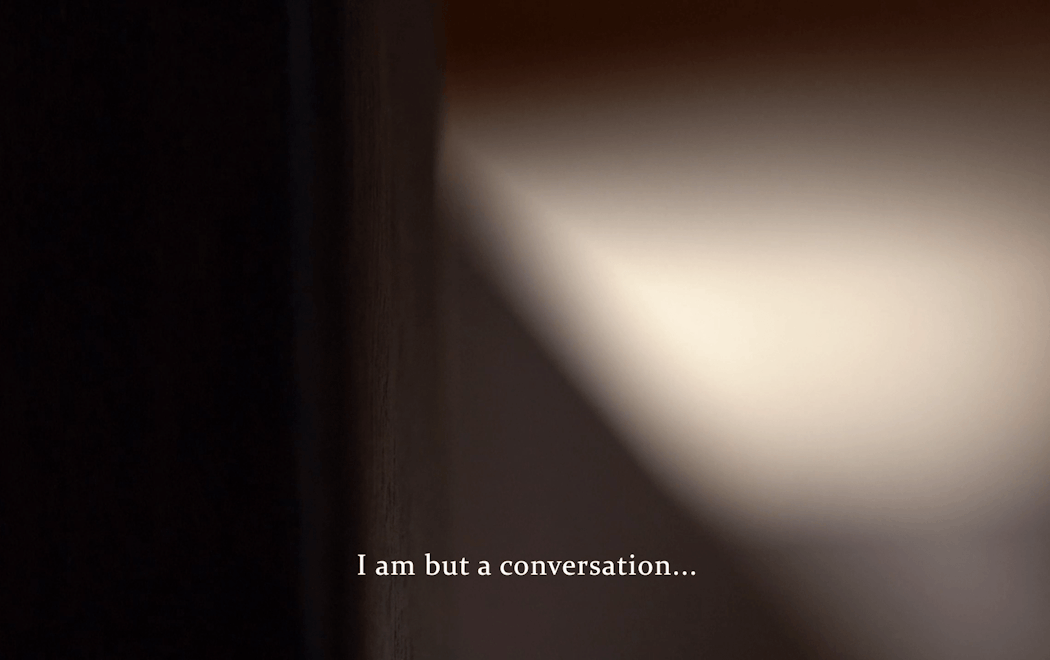 An indistinct blur of white light next to a block of black. At the bottom of the screen is a caption which reads "I am but a conversation..."