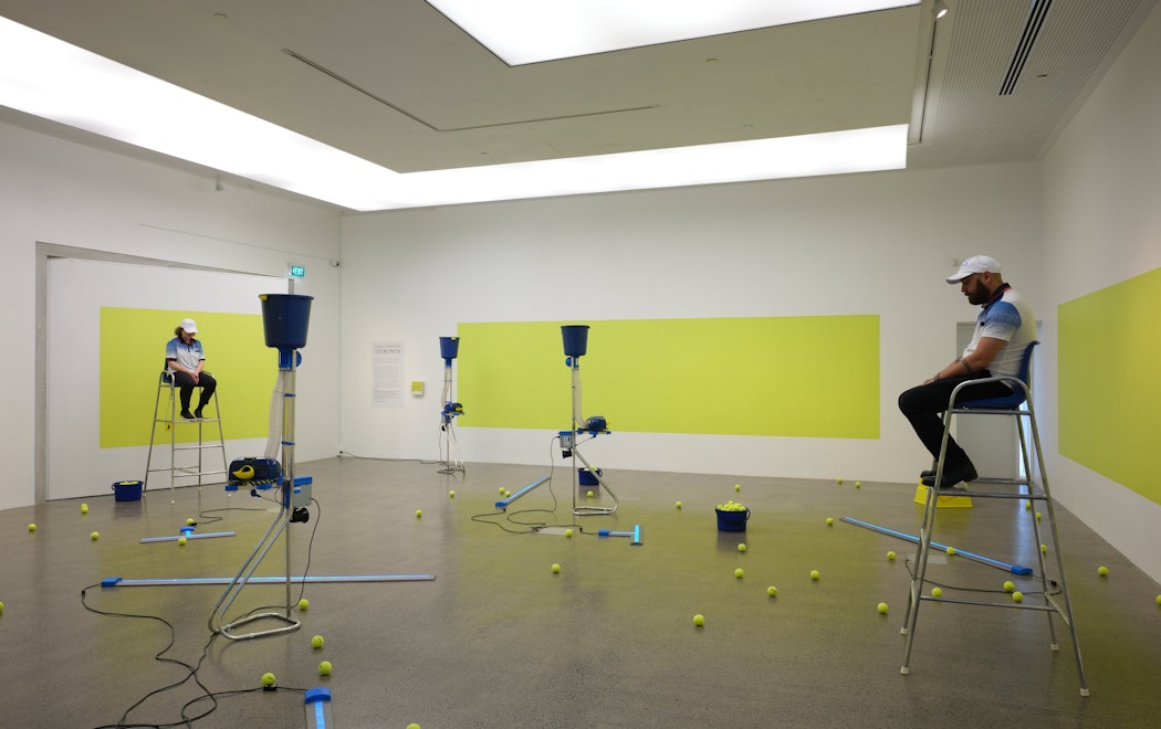 Two people sit on umpire chairs on either side of a brightly lit gallery. Sprawled upon the gallery floor are tennis ball shooter machines, tennis balls are dotted throughout then space.