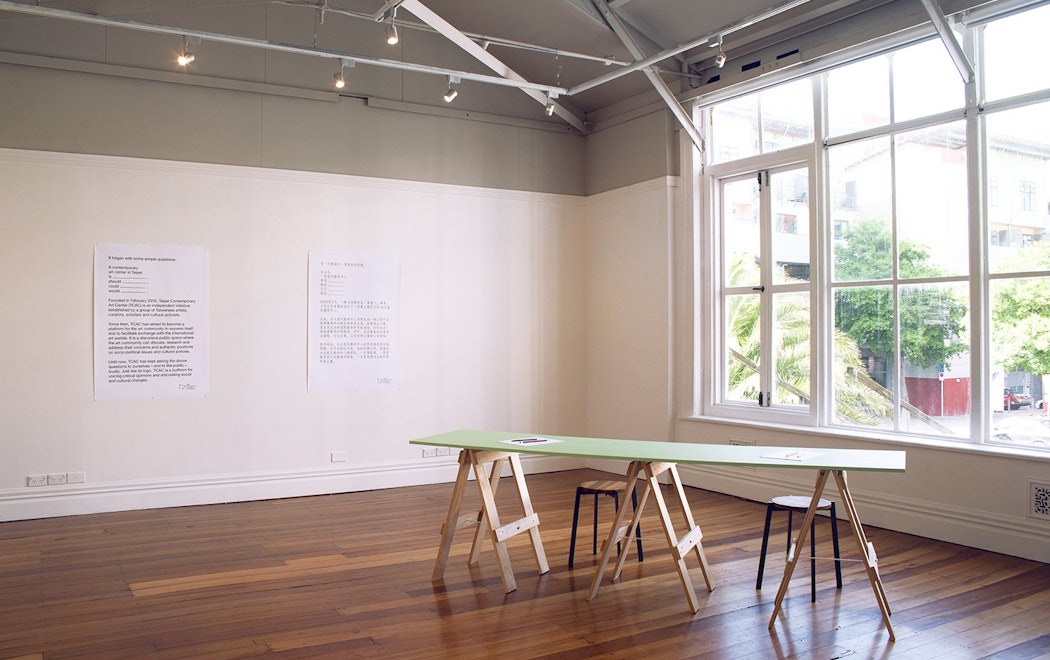Bright light streams through large windows into a gallery with woooden floors, two large text images are on the wall and there is a table in the foreground