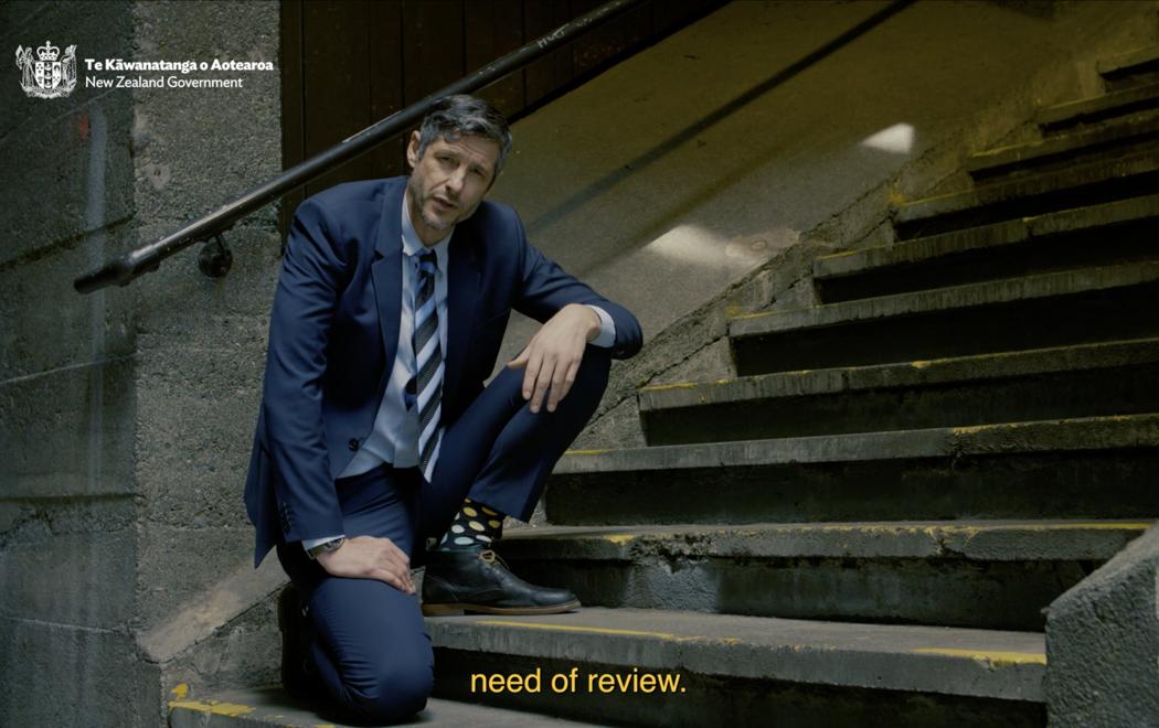 A man in a blue suit and tie kneels on a concrete staircase facing the camera, as if to give an explanation. At top left we see a logo for the New Zealand government. On the screen, we see the words 'need of review'. The man's colourful socks cast doubt on his apparent authority and point towards satire.