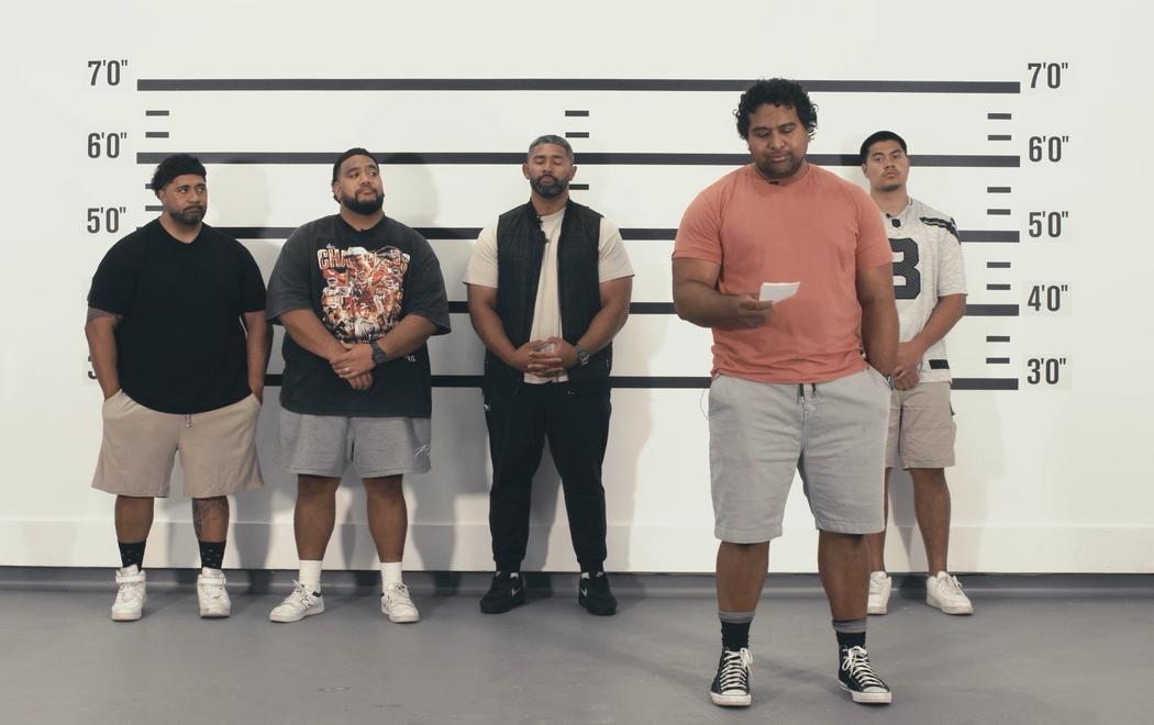 Five Pasifika males in casual clothing line up against a wall as if in a police line up. One is stepping forward to read from a note in their hand.