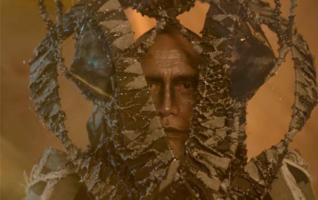 The face of a male figure shows a stern expression. Their head is enveloped by a series of green wavy tendrils, which point upright.