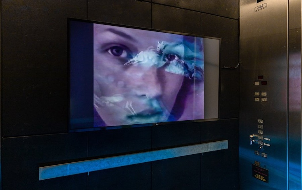 Inside a lift, screen showing Kate Moss's face with animated figures overlaying it.