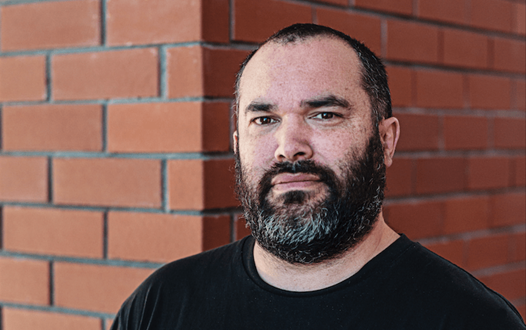 Rangituhia wears a black t-shirt and has a dark beard, he looks at the camera with a serious expression