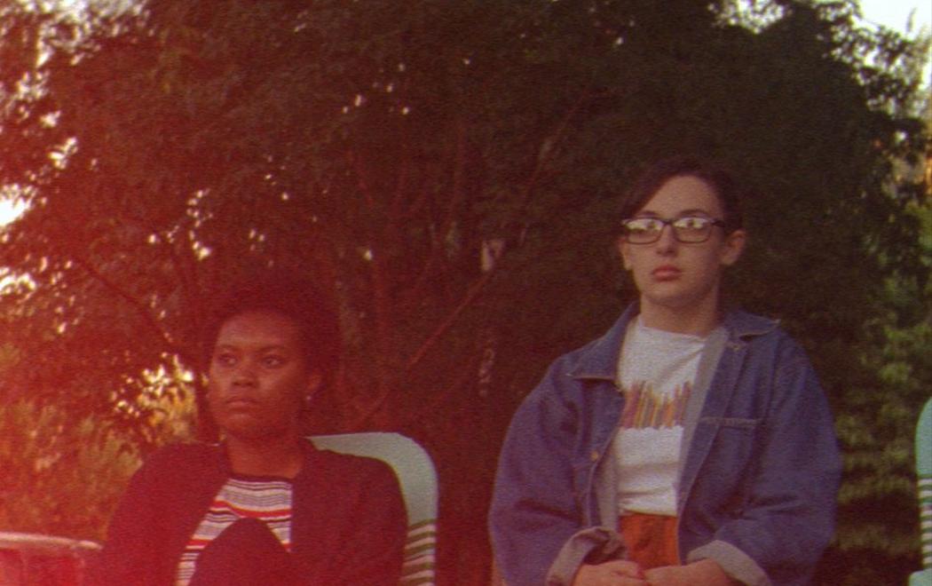 Teenagers sit on deck chairs on the pavement looking out into the distance with bored expressions