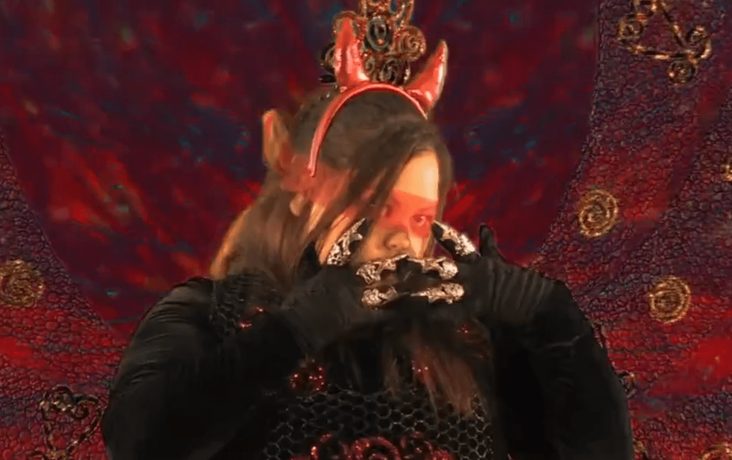 Candice is dressed in glamorous costuming, wearing black gloves with jewellery on top, devil horns and a red and black dress. They are dancing in front of a swirling red textured background