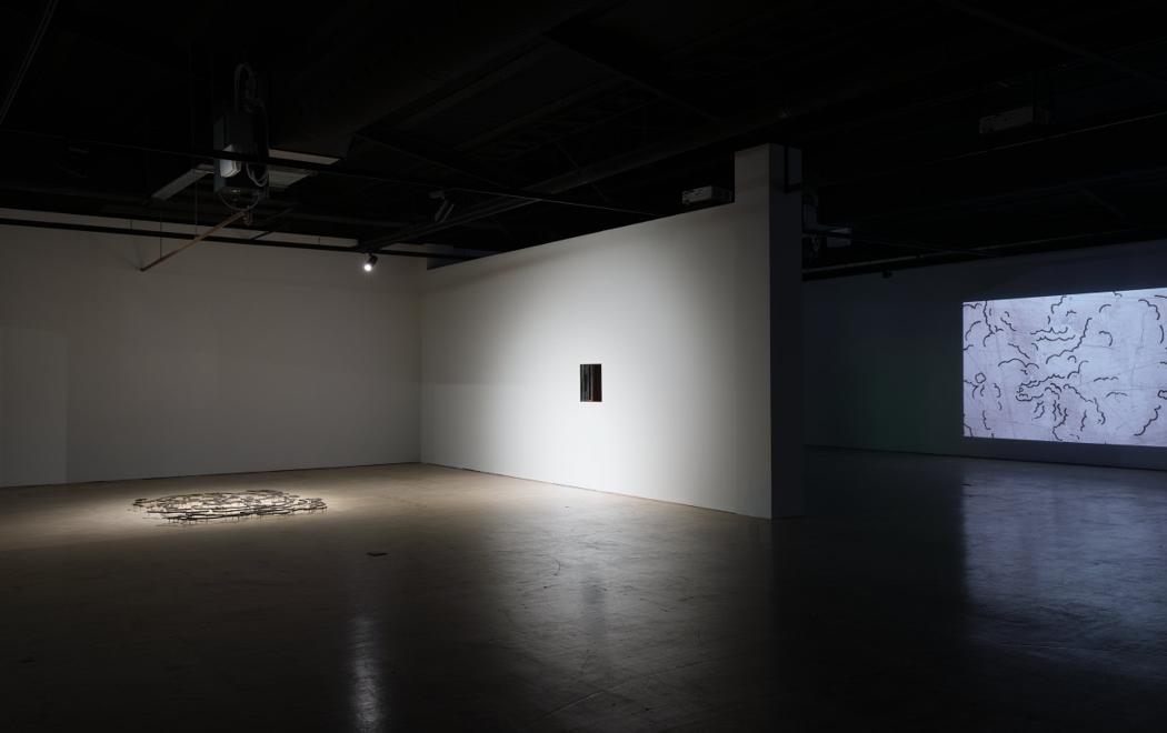 In a gallery we see three artowrks, from left, on the floor a loose assembly of metal resembling a cloud, in the centre a hole in the wall approximately the size of a dumb waiter, and at right, a video projection of an animated cloud.