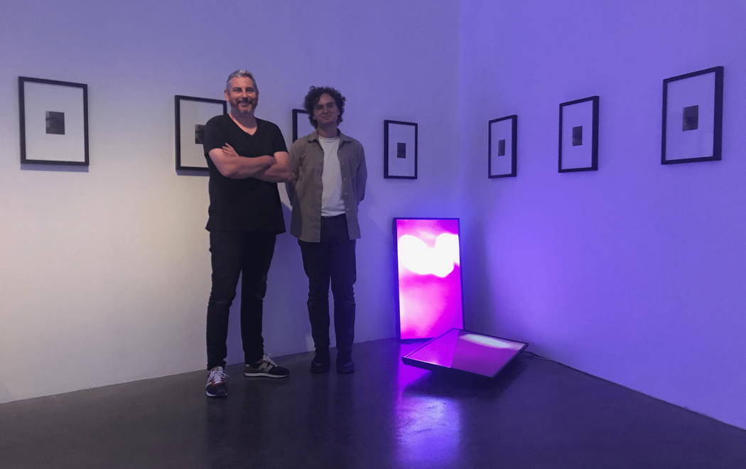 Steve and Christian stand in a gallery which displays small photographs in large frames neatly on the walls, two tvs rest against the wall and the floor of the gallery displaying a bright, pink image