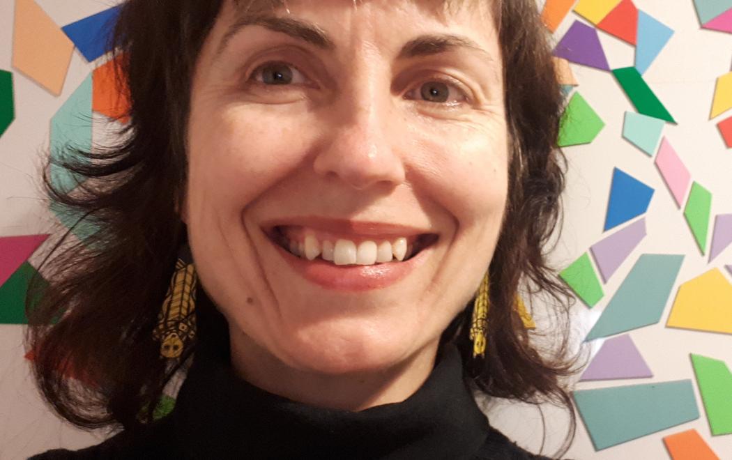 A woman smiles for the camera, behind her are colourful mosaic tiles stuck to the wall.