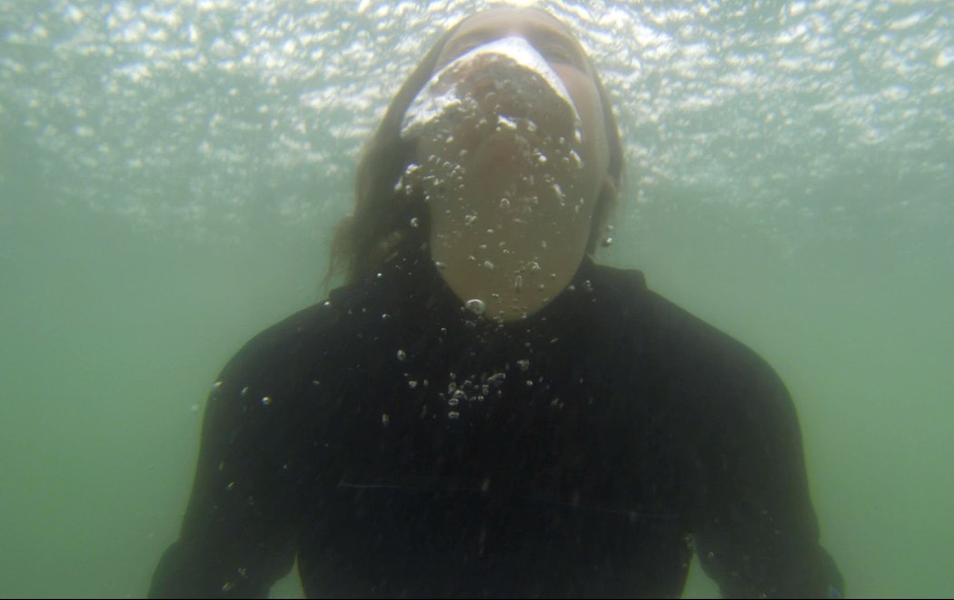 Alex is underwater wearing a wetsuit in a green ocean. She is coming up for air and is exhaling creating large bubbles in the water