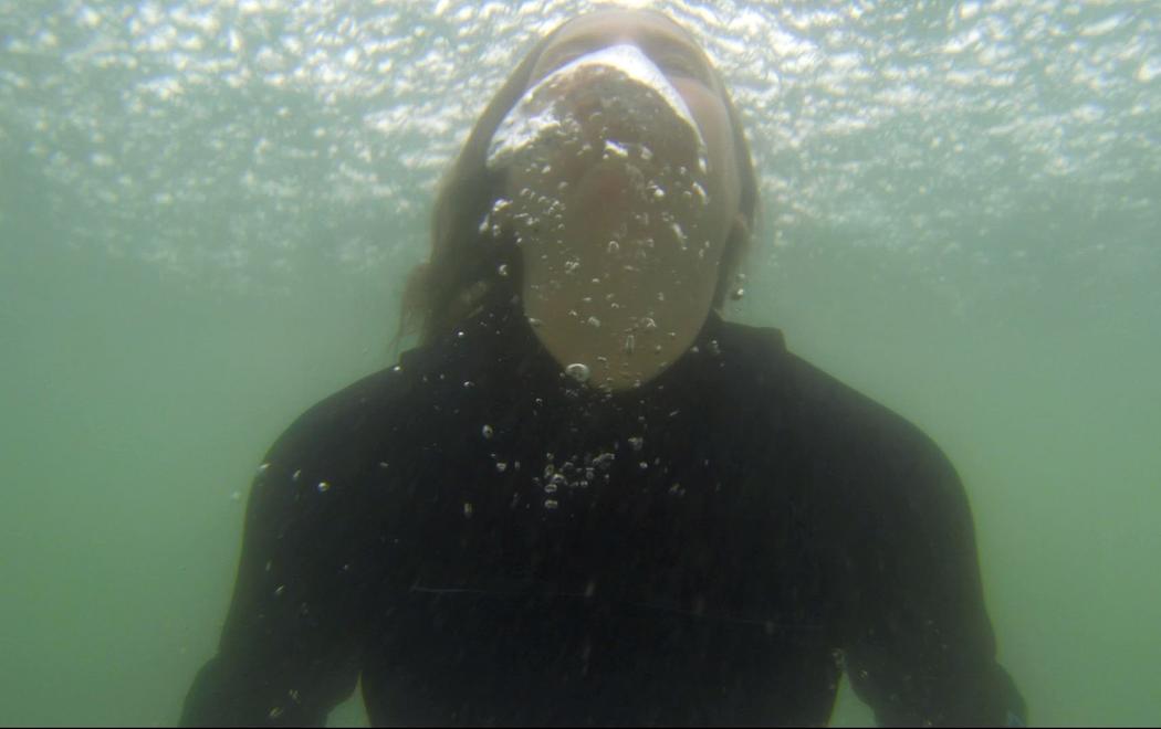 Alex is underwater wearing a wetsuit in a green ocean. She is coming up for air and is exhaling creating large bubbles in the water