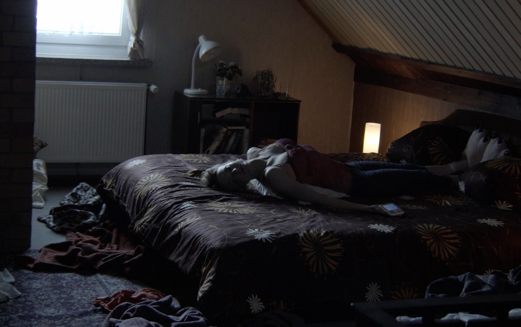 In a messy, dimly-lit bedroom, a women lies on the bed with her back arched and feet on the pillows.