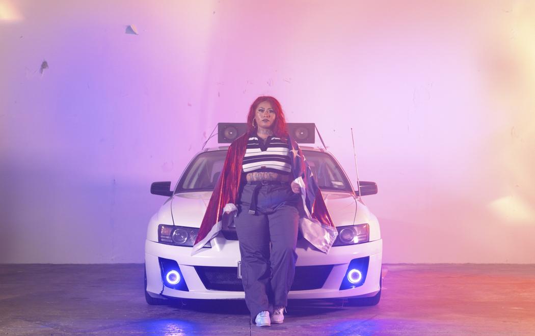 A young Pasifika woman leans steadfastly against the bonnet of a boy racer style car, which has speakers mounted on the roof