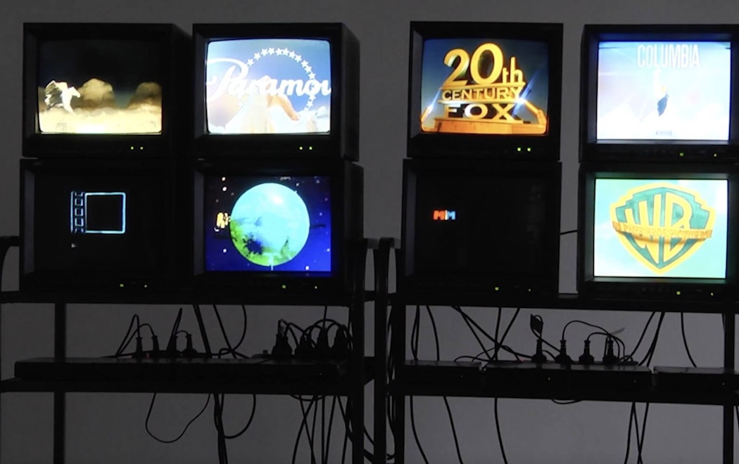 Eight vintage TV monitors are stacked in 2 x 2 configurations and displayed on black shelves with numerous wires and power cords visible. Each monitor shows a Hollywood movie studio logo.