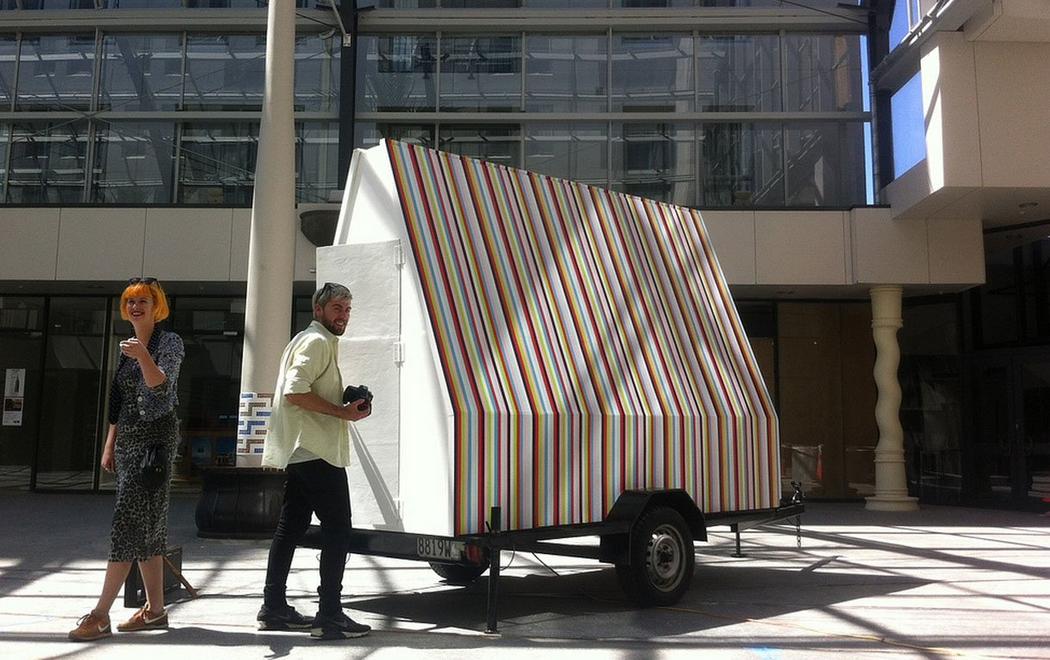 Two people stand next to a large house shaped object which is sitting on a trailer. The house or object is large enough that a person could potentially go inside, it is painted thin precise stripes or black, red, green, pink, blue and baby blue. The two people standing next to the object look happy and excited in the sun coming in from the windows.