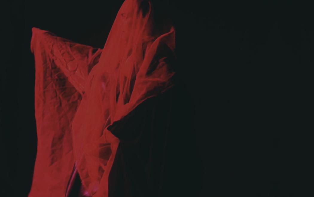 A figure is covered with sheer fabric and awash with red light against a black background