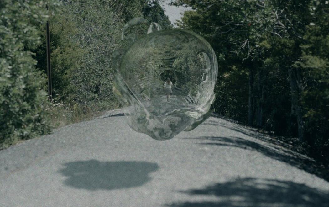 A large water bubble looms over a rural bush road, in the distance a female figure walks with her back to us