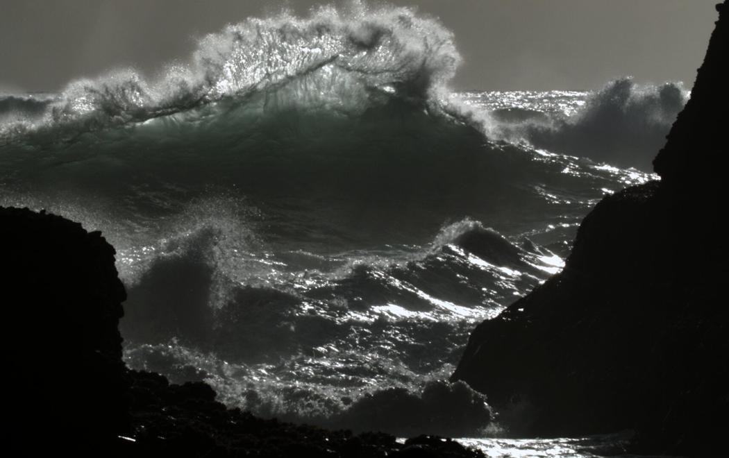 A stormy sea with a large wave breaking in the middle and rocks seen close by.