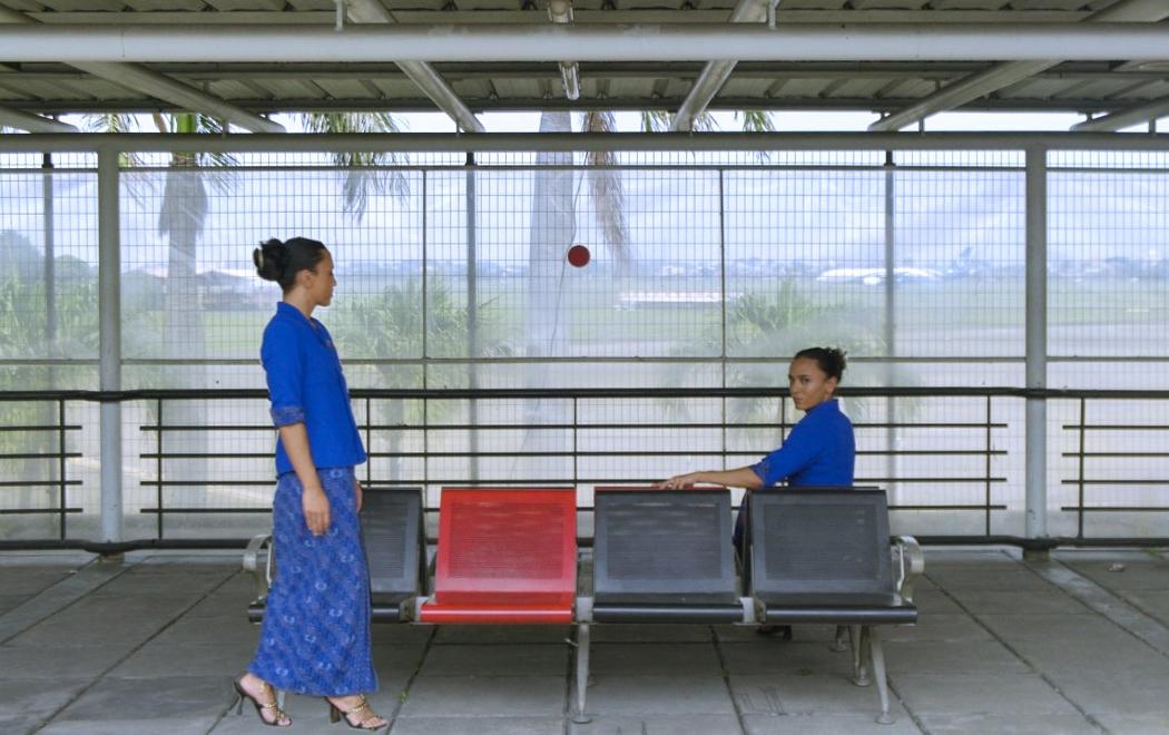Two women in kebaya dress stand and sit next to an outdoor bench at an airport