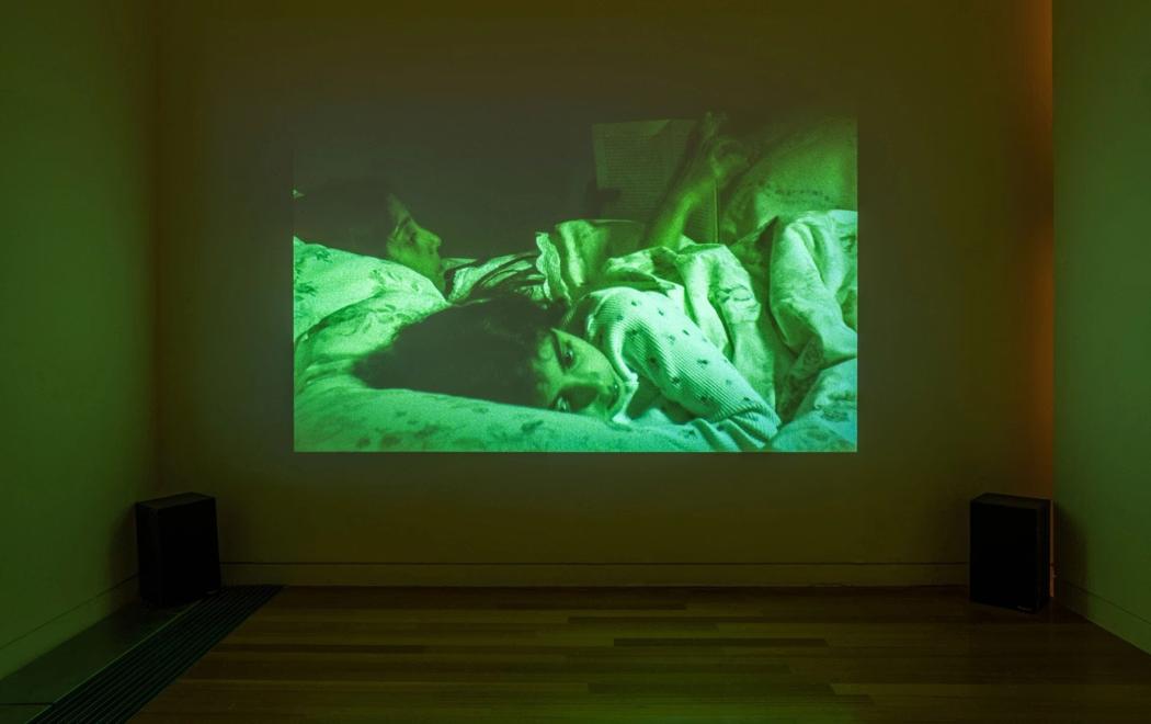In a gallery we see a projected image of two young people in bed, each of whom is awake at night. The image is cast in a green luminescent hue.