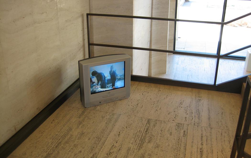 A small video monitor sits on the floor in the corner of what appears to be a stairwell. The monitor screen is in the early television ratio of 4:3, as opposed to the contemporary landscape format of 16:9. The monitor is an installation of Julian Dashper's video work Studio Songs (1998), installed at Sheldon Museum, Lincoln, Nebraska (2006).