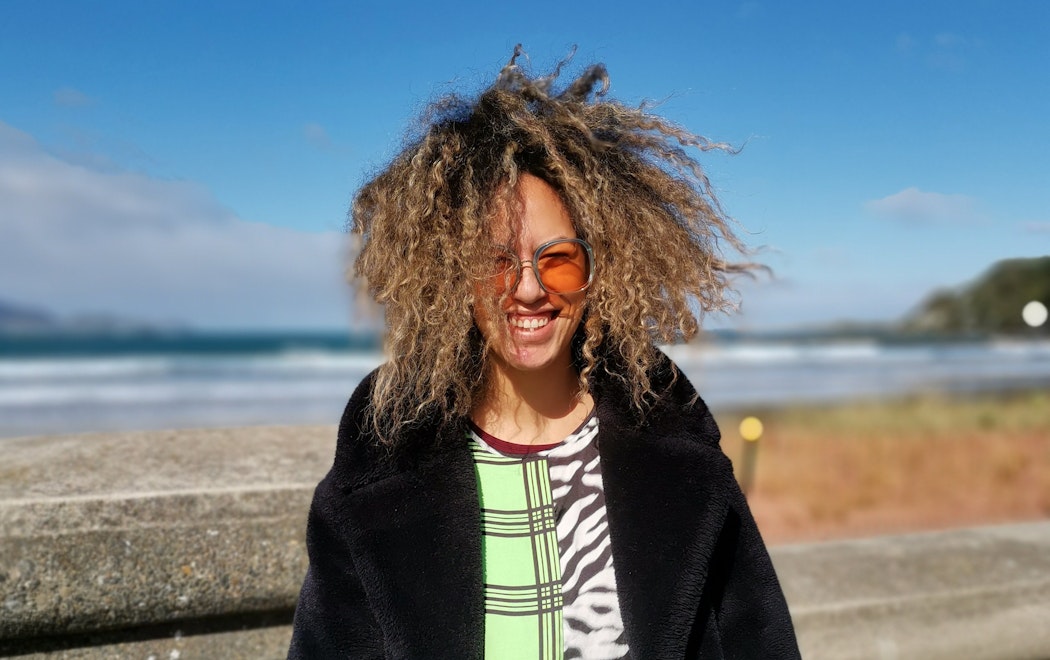 A photo of Israel Randell standing facing the camera. She wears sunglasses and her curly hair is blown by the wind. Behind her is the ocean.