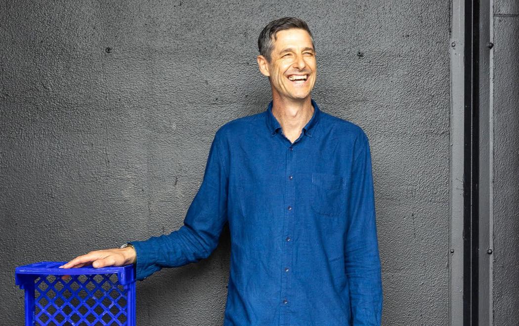 A grinning man in a blue shirt stands against a grey wall; his hand is resting on a stack of blue plastic crates.