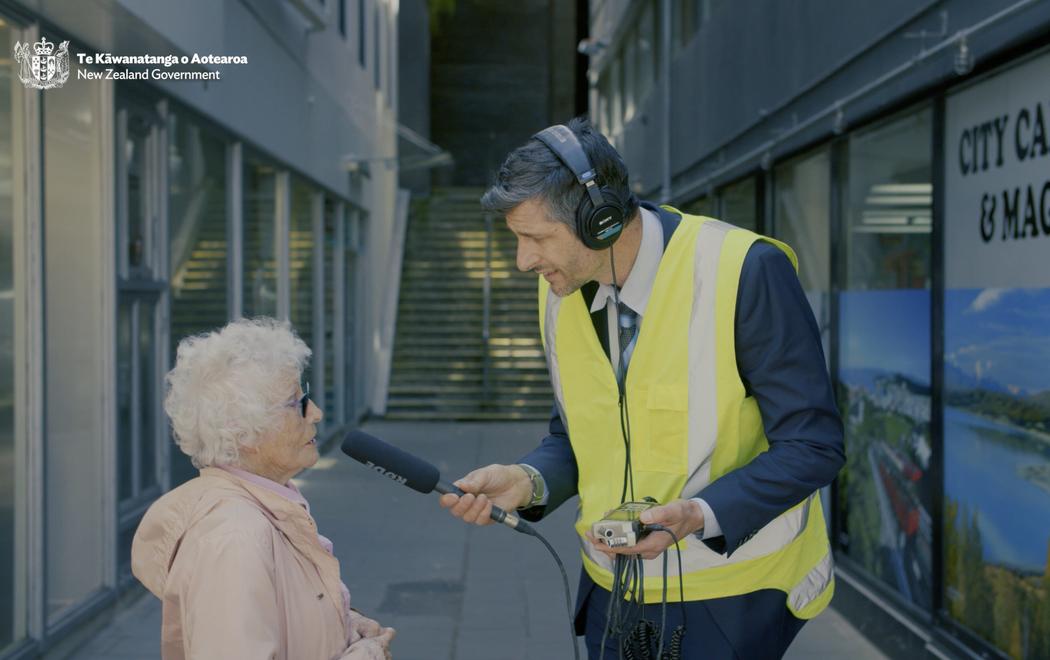 A tall man in a suit and high visibility vest interviews a comparitively dimunutive old lady on the street.