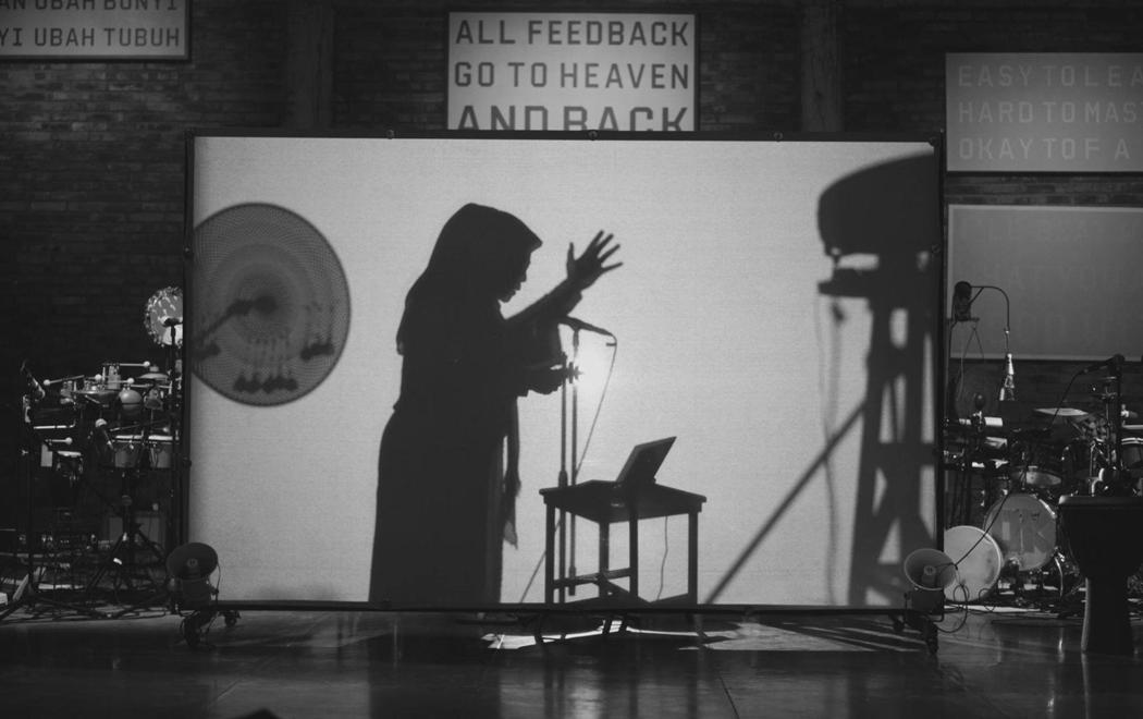 Behind a screen we see the shadow of a woman gesturing upwards whilst speaking into a microphone. At either side of the screen are drums and percussion. On the wall are texts in Indonesian and English. Above the screen reads "ALL FEEDBACK GO TO HEAVEN AND BACK".