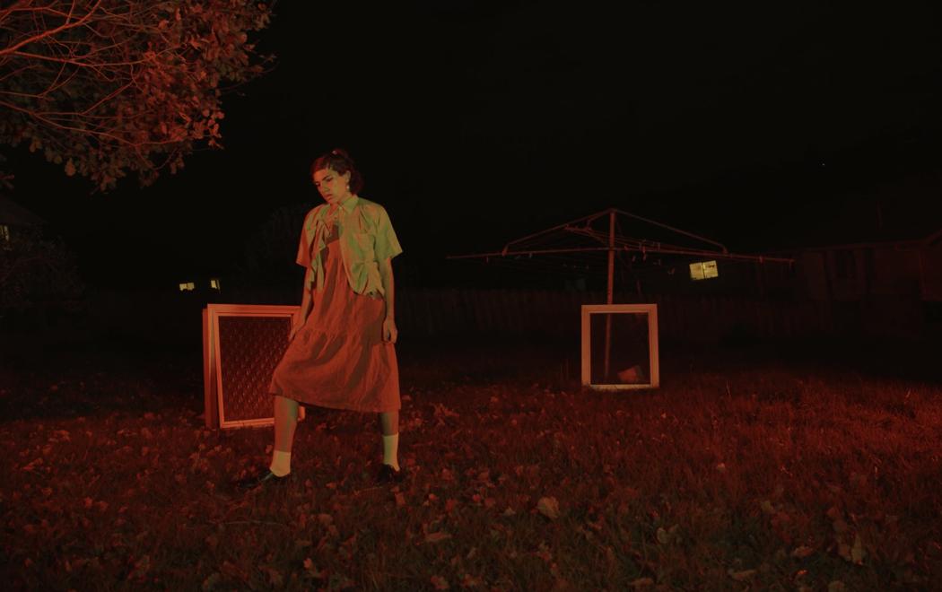 In a backyard a person in a dress stands in front of two windows which are propped up on the lawn. The second window rests against a clothes line. The scene is lit by a red glow.