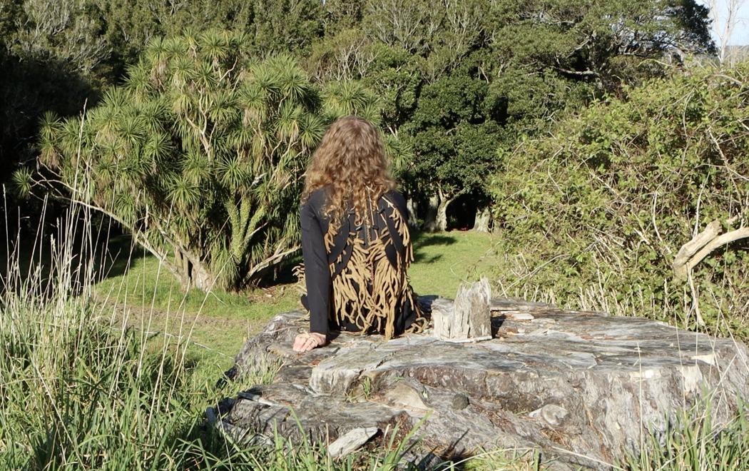A person sits on a tree stump with their back to camera, they face trees and the sun is shining. They wear adornment made partly from tree leaves.