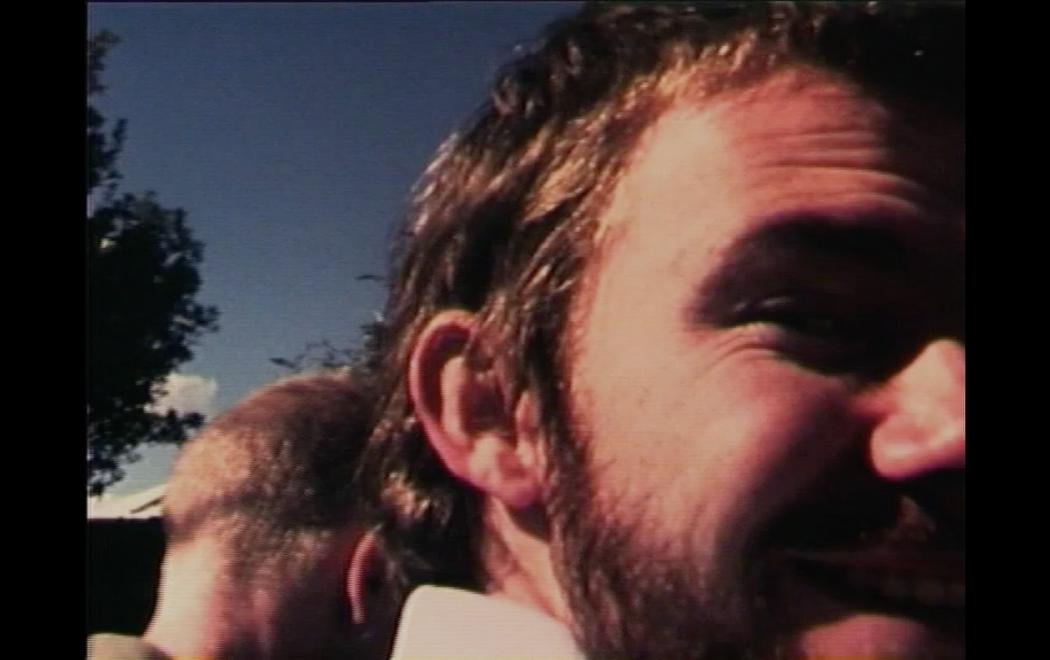 A man's face turns and smiles to the camera; another man's head is visible behind him