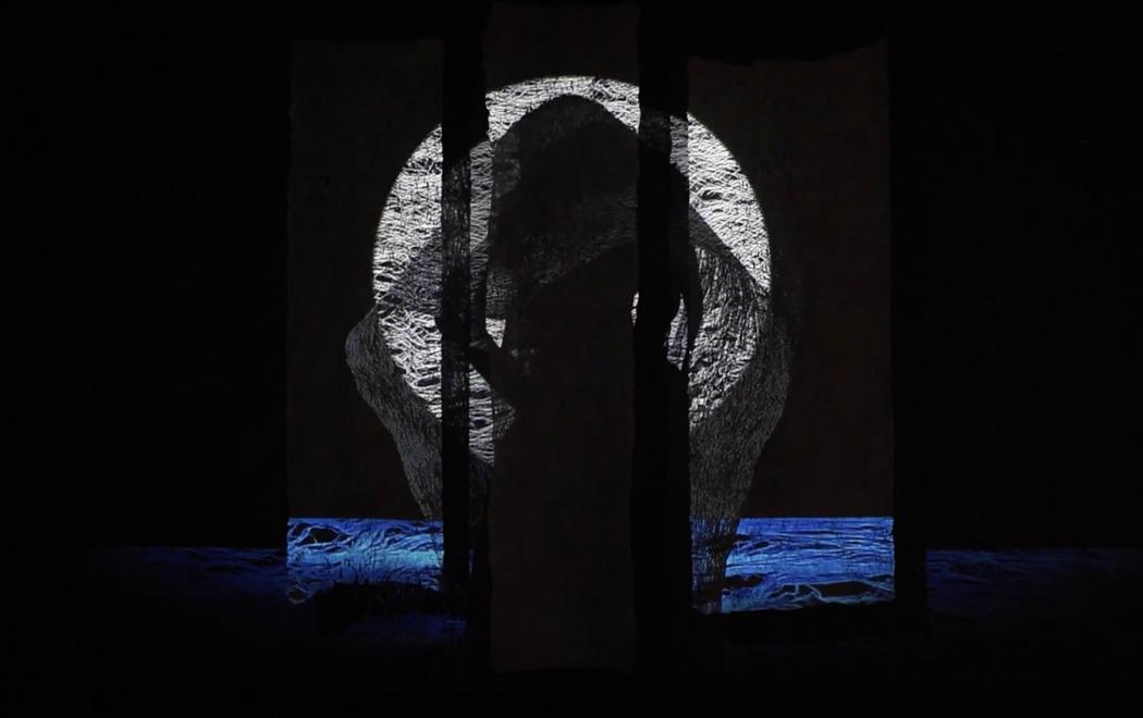 A white circular form is projected across three panels of barkcloth, while a blue band resembling water extends along the bottom edge of the projection. The silhouette of a figure draped in tapa lies at the centre.