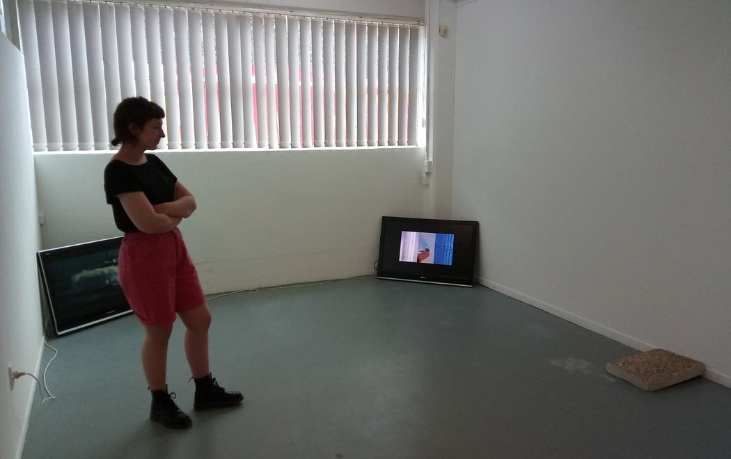 Priscilla Howe, CIRCUIT's 2017 Curatorial Intern, stands in The Audio Foundation gallery looking at an installation of artworks. The works include two videos on separate monitors and a slab of concrete. This is the installation of the exhibition PULSE/REPEAT, a grooup show curated by Priscilla Howe at The Audio Foundation, Auckland (2017).