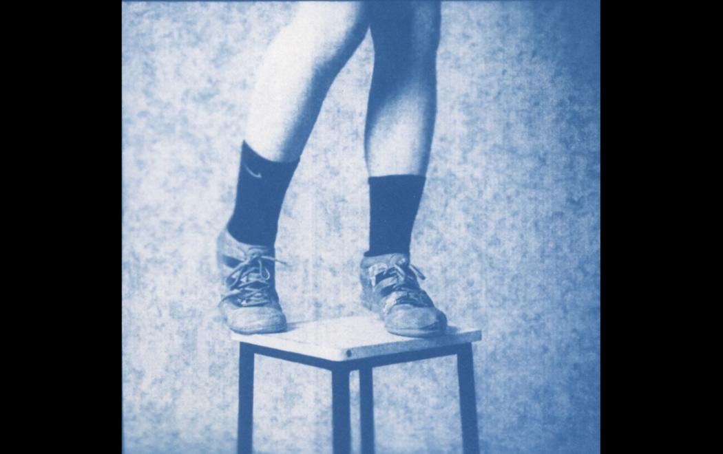 A sepia toned image of a person's lower body on a stool. We see their bare legs, socks, and shoes. Their feet are apart as if balanced, one is curled as if to balance on the toes.The background is a flat, textured surface, as if a domestic space or a photographic studio. The stool is a plain white functional square on black metal legs.