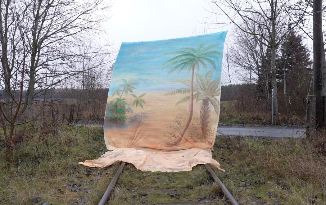 A painted background of palm trees, sand and sea is hung over a railroad track in the middle of what appears to be rural Europe. The sky is grey. The railway track points towards a road, indicating the track is now unused.