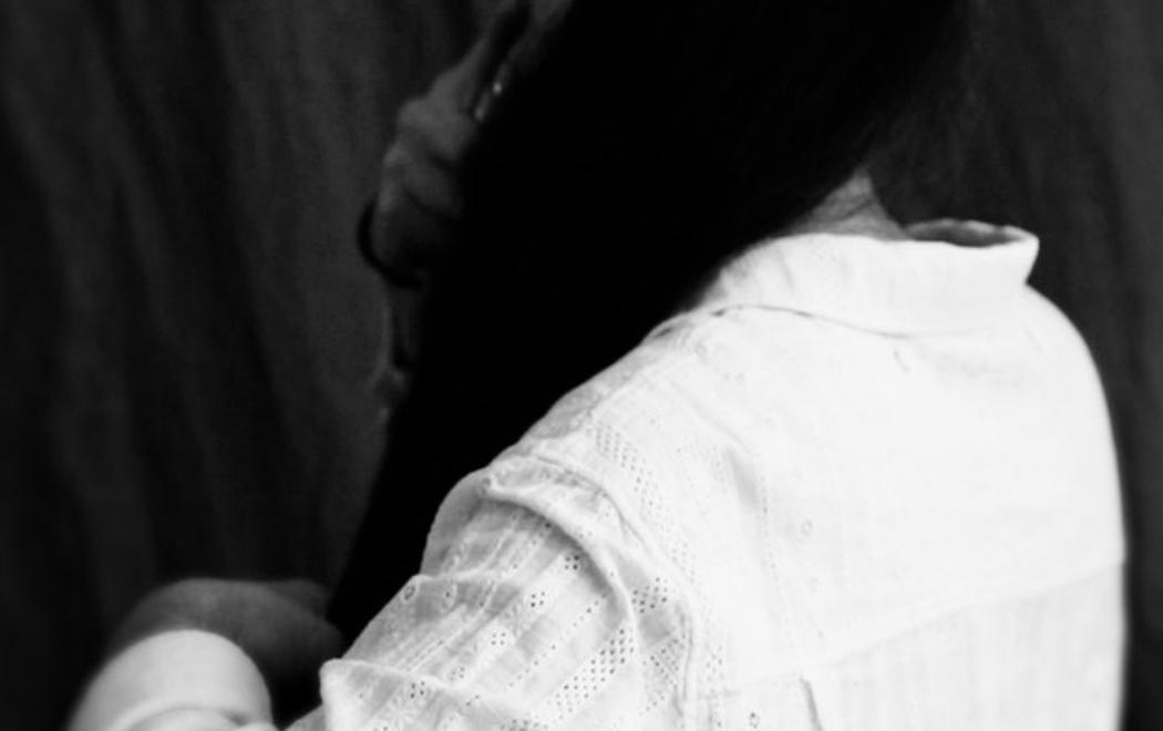 A black and white portrait of a person brushing their hair. They face away from the camera, their top is a white linen short or blouse. The image suggests a portraiture tradition from the Colonial era.