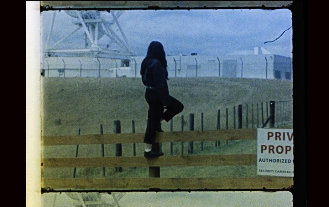 A person stands on a rural fence looking at an astronomy observatory behind a wire security fence