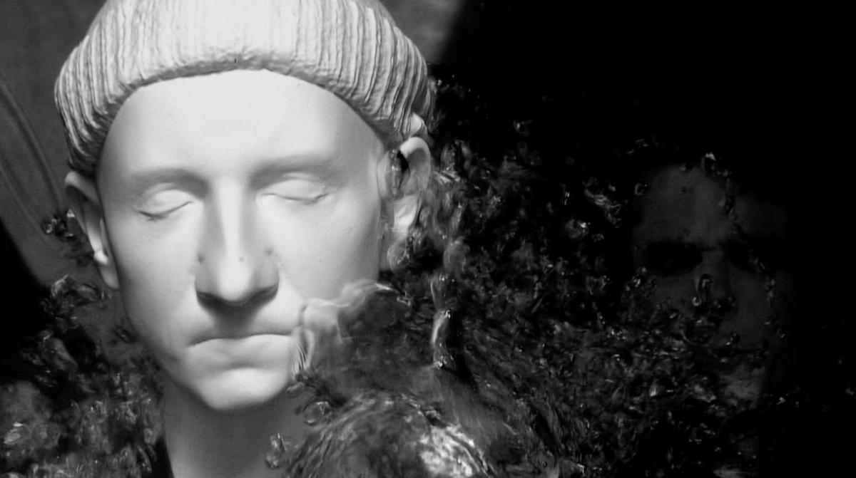 A computer generated model of the artists face is being splashed by water. The face has eyes closed and wears a beanie. It has been rendered white.