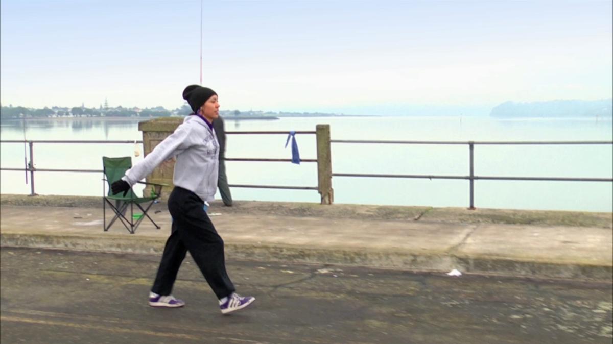 A person in a tracksuit and beanie dances along a bridge.