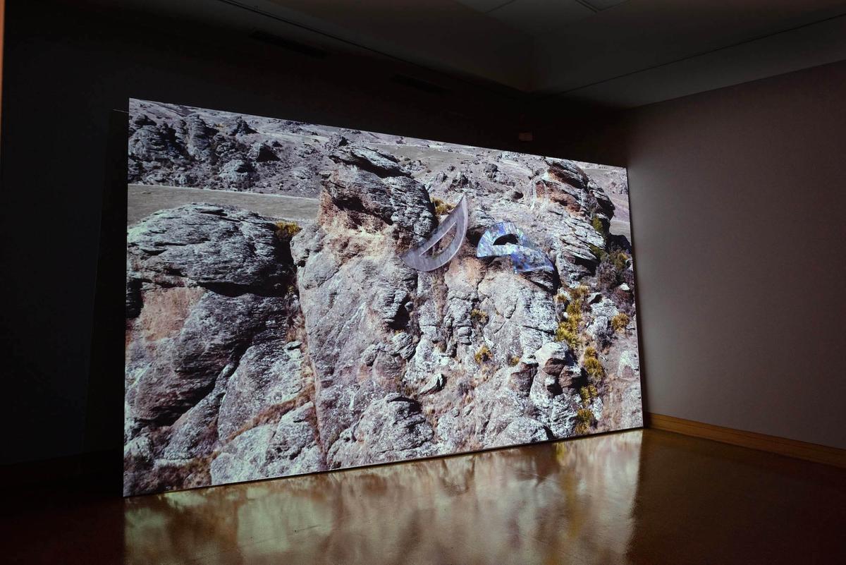 A titled film screen in a darkened room, the image shows a panoramic view of rocky hills overlaid with digitally rendered half moon shapes.