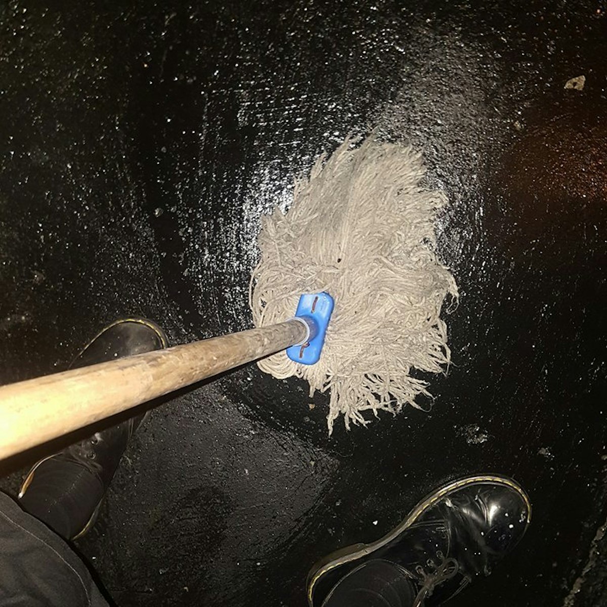 A hairy mop is being pushed against a shiny black concrete floor in a sex on site club. The image is taken looking down at the mop and the artists feet, mid way through mopping the floor.
