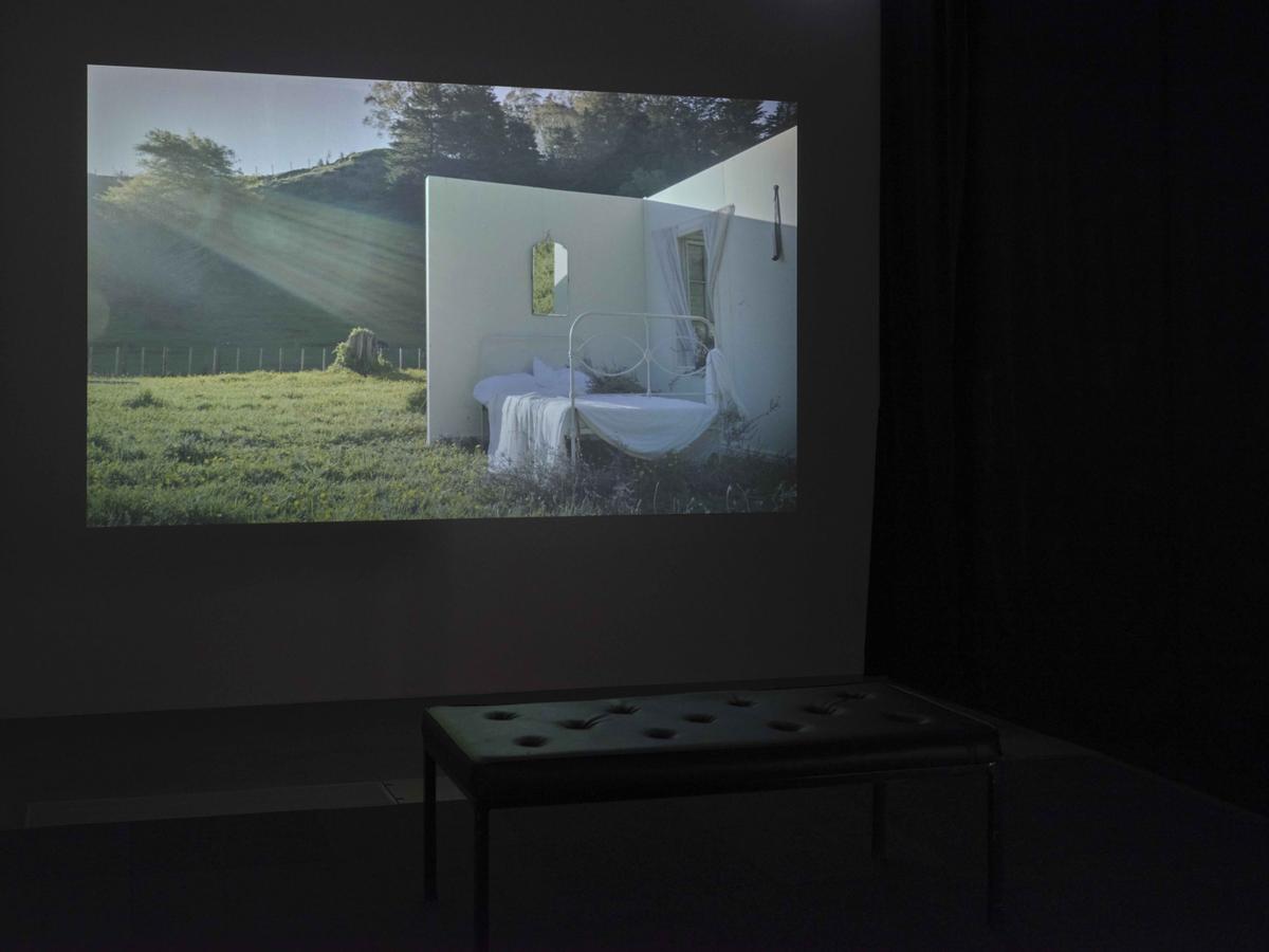 Sunlight streams into an installation on a grassy field which looks like a film set of a scene. We see an unmade bed with white linen against the only two existing walls