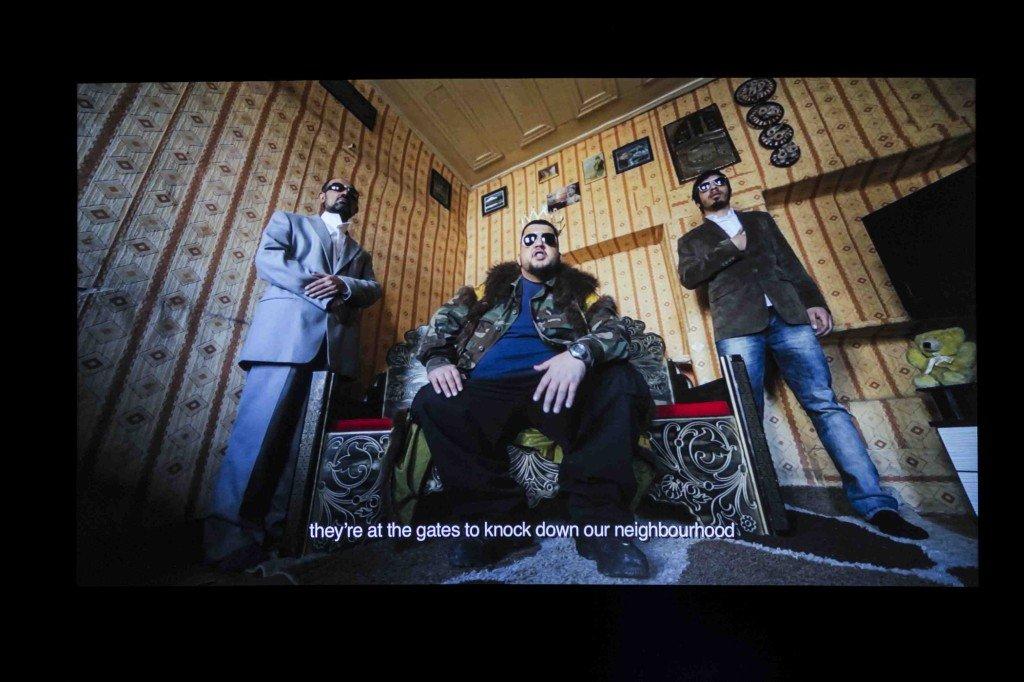 A wide angle shot taken from low down looking up of three dapper looking rappers in a domestic setting has the words "they're at the gates to knock down our neighbourhgood"