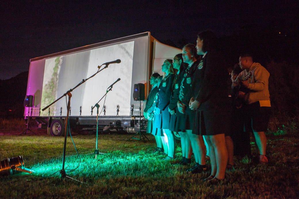 A group og high school students sing in front of an image projected on a truck