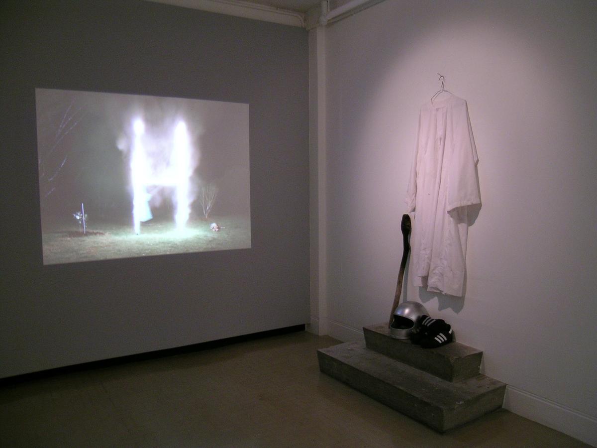 In a gallery a video projection shows a set of rugby goalposts on fire, while at right a white robe hangs on the wall. Below it are a silver motorcycle crash helmet, a pair of adidas sneakers and a wooden staff with a charred end.